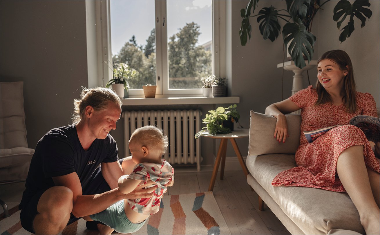 Nadja Huotari puolisonsa ja lapsensa kanssa olohuoneessa, Nadja istuu sohvalla ja lapsi ja puoliso ovat lattialla.